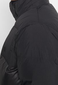 Svart puffjacka med hög krage, med en slät ytteryta, stickade sömmar och en logotyp på axeln.