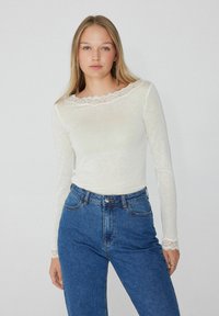 Weißes Langarmshirt mit Spitze am Ausschnitt und an den Bündchen, kombiniert mit hochgeschnittenen blauen Jeans. Leichter, strukturierter Stoff.