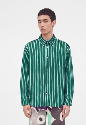 Young man wearing a green and white vertical striped button-up shirt and pants with large abstract floral patterns, standing against a plain background.