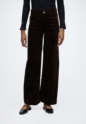 Pantalon large en velours côtelé marron foncé avec bouton et fermeture éclair à l'avant, porté avec un haut à manches longues noir côtelé et des chaussures Mary Jane noires.