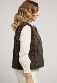 Gilet en fausse fourrure de couleur gris foncé, avec une coupe décontractée, sans manches et doté de poches latérales. Porté par-dessus une chemise à manches longues blanche.