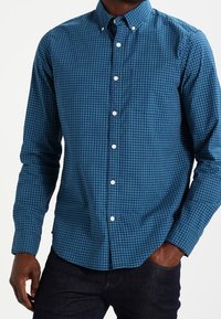 Chemise boutonnée à carreaux bleus avec col large, manches longues et boutons blancs. Le tissu a une texture lisse avec une coupe régulière.