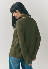 Olive green long-sleeve blouse with a soft texture and a high collar, featuring open back detail and loose fit, paired with blue jeans.