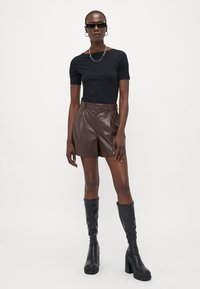 Haut noir ajusté, shorts en cuir marron taille haute, et bottes compensées noires à mi-mollet. Les accessoires comprennent des lunettes de soleil et un collier en chaîne argenté.