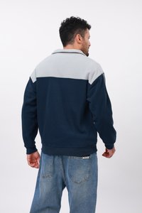 Uomo che indossa una giacca in fleece bicolore con la parte superiore grigia e la parte inferiore blu navy, abbinata a jeans blu larghi, di spalle contro uno sfondo neutro.