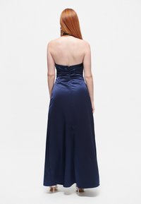 Pitkä, tummansininen satiini-ilta-asu, jossa on istuva bodice ja virtaava hame. Takana on kokoontunutta yksityiskohtaa ja olkaimeton muotoilu.