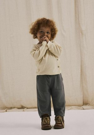 Bambino che indossa un maglione a maglia color crema con bottoni e pantaloni grigi tapered, accompagnati da stivaletti marroni con lacci. Tessuto texture e vestibilità loose.