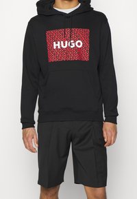 Černá mikina s kapucí a červeným vzorovaným potiskem, na kterém je bílým písmem napsáno "HUGO", v kombinaci s černými šortkami. Vyrobeno z jemného materiálu.