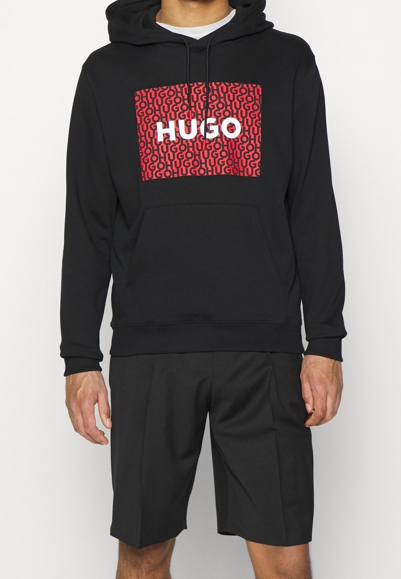 Černá mikina s kapucí a červeným vzorovaným potiskem, na kterém je bílým písmem napsáno "HUGO", v kombinaci s černými šortkami. Vyrobeno z jemného materiálu.