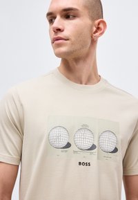 Fiatal férfi bézs pólóban, amelyen három golflabda műszaki rajza látható, alatta a "BOSS" felirattal, balra néz.