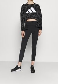 Svart atletisk kort sweatshirt med vit Adidas-logotyp, kombinerad med högmidjade svarta leggings med vita sidostråk och svarta sneakers.