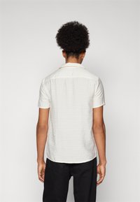 Koszula z krótkim rękawem w teksturowanym off-white materiale w poziome paski, z kołnierzykiem i zaokrąglonym dołem, noszona z ciemnymi spodniami.