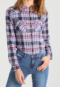 Rutigt flanellskjorta i rött, marinblått och vitt med klassisk krage, två bröstfickor och knäppningar. Bärs med blåjeans.