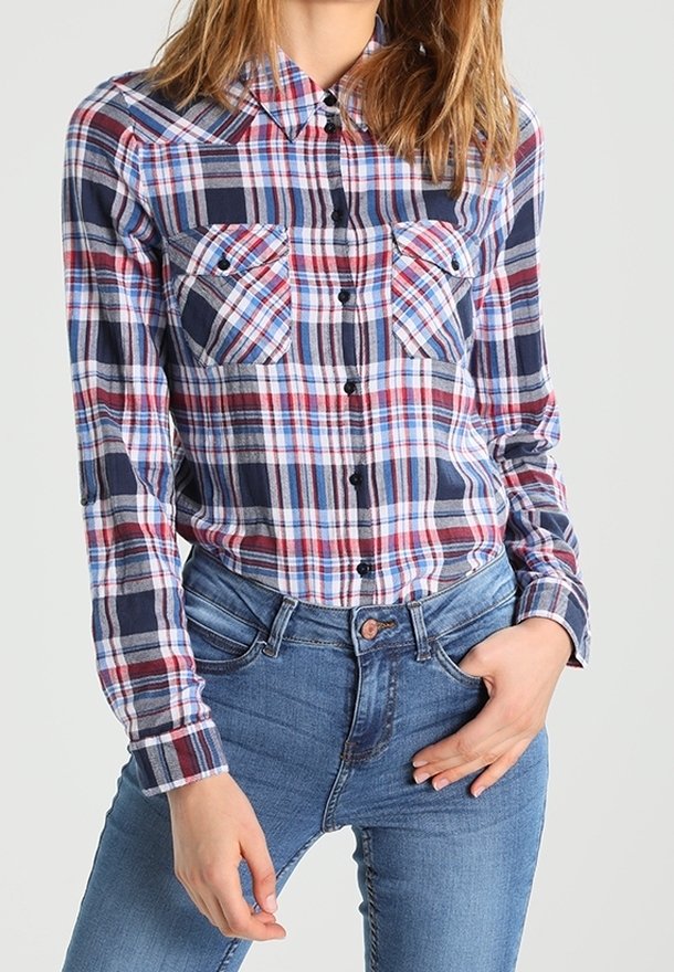 Rutigt flanellskjorta i rött, marinblått och vitt med klassisk krage, två bröstfickor och knäppningar. Bärs med blåjeans.