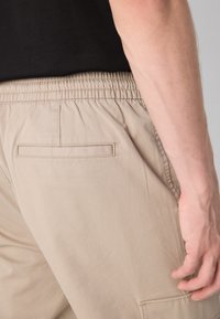 Beige elastische taillebroek met een achterzak, een gladde stofstructuur en een getailleerde snit, zijaanzicht weergegeven, gecombineerd met een zwart shirt.