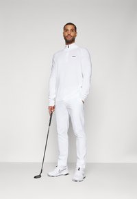 Bela golfska oprema, ki vključuje dolg rokav s cevjo na vratu in logotipom, usklajene hlače in bele golf čevlje. Drži golf palico.