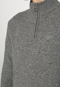 Gros plan sur une personne portant un pull gris texturé en tricot avec une fermeture éclair demi-fermeture et un petit logo Barbour brodé sur la poitrine.