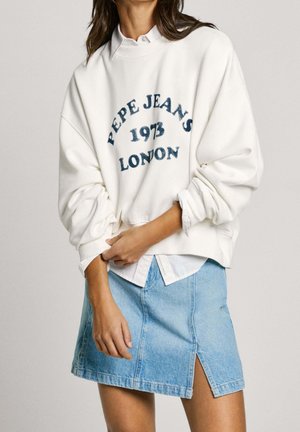 Nainen pukeutuneena valkoiseen Pepe Jeans -collegepaitaan, jossa teksti "1973 London", valkoisen paidan päällä, yhdistetty vaaleansiniseen farkkuhameeseen, jossa on halkio edessä.