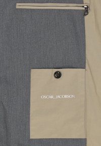 Tessuto grigio con una texture liscia presenta una tasca beige con un bottone nero e il logo "OSCAR JACOBSON" ricamato in bianco. Tasca con zip sopra.
