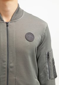 Giacca bomber verde oliva con zip frontale, colletto a coste e una patch rotonda nera con logo. Presenta una tasca sulla manica con zip e un tessuto testurizzato.