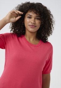T-shirt s krátkym rukávom a okrúhlym výstrihom v žiarivej koralovej farbe, vyrobený z mäkkej látky s hladkým povrchom. Vyznačuje sa voľným strihom a minimálnym počtom švíkov.