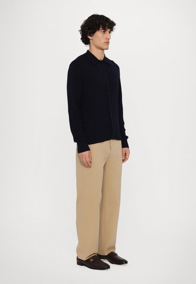 Cardigan bleumarin texturat cu nasturi, asortat cu pantaloni bej și mocasini maro, stând în fața unui fundal simplu.