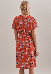 Röd klänning med korta ärmar, med ett färgglatt fågelmönster. Designen inkluderar en figursydd midja och en liten nyckelhålsöppning i ryggen.