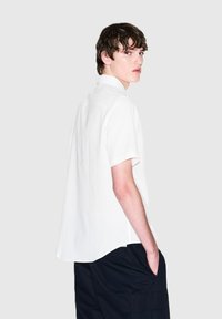 Camicetta bianca a maniche corte con un design testurizzato, dotata di un colletto classico e una parte posteriore leggermente più lunga, abbinata a pantaloni scuri.