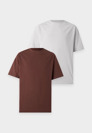 Deux jeunes adultes debout côte à côte portant des t-shirts oversize gris clair, des pantalons noirs à jambes larges et des baskets noires sur fond blanc.