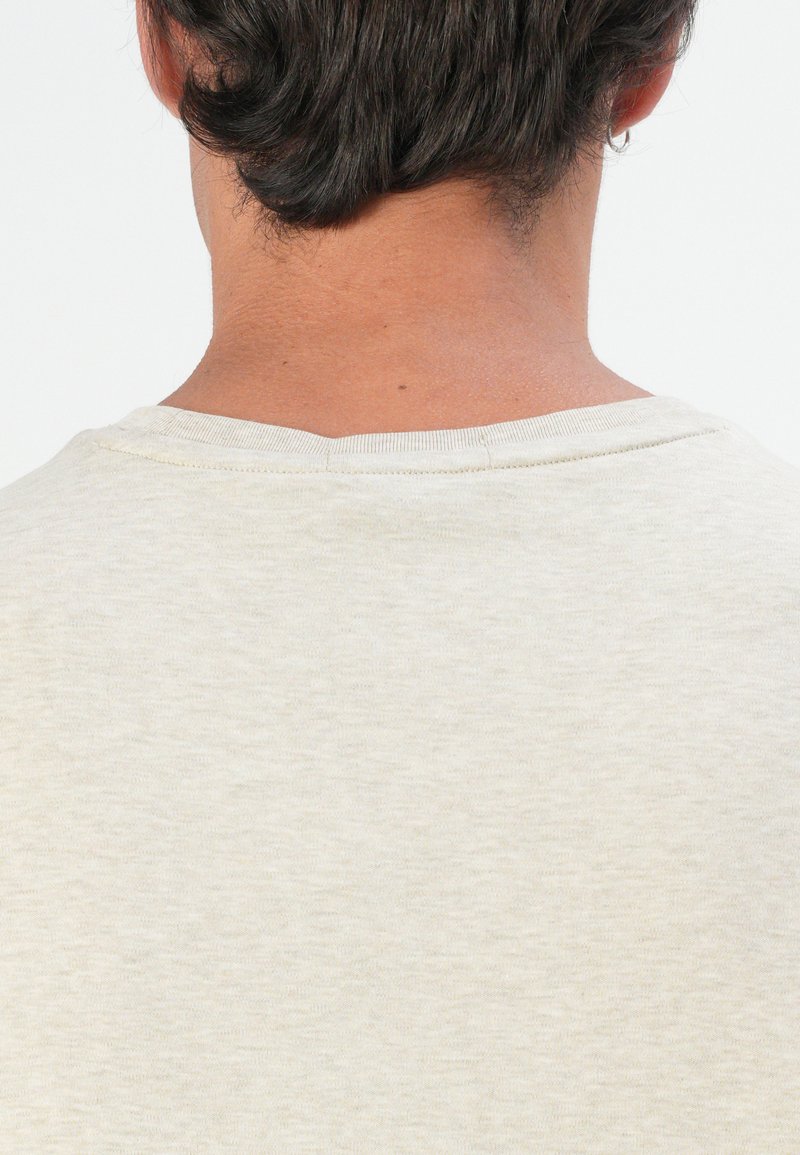 Camiseta de color beige claro con cuello redondo, textura suave y un diseño sencillo. La tela parece ligeramente jaspeada. Vista trasera únicamente.