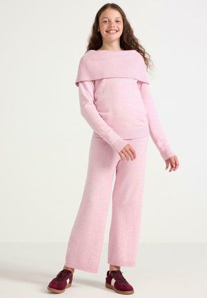 Lächelndes Mädchen mit langen lockigen Haaren, das einen rosa gestrickten Pullover und eine Hose trägt und in einem neutralen Raum mit bordeauxroten Sneakers steht.