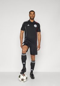 Zwart voetbaltenue bestaande uit een poloshirt met witte en rode accenten, gecombineerd met shorts en kniekousen. Bal aan de voet. Adidas merk.