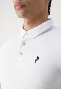 Vit polo skjorta av texturerad tyg med en tre-knappslå och en svart logotypsaccent på vänster sida.