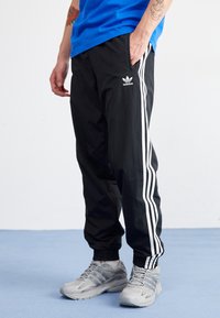 Svarta träningsbyxor med vita sidostrikar, elastiska muddarna och en Adidas-logotyp. Bärs med en blå skjorta och grå sneakers.