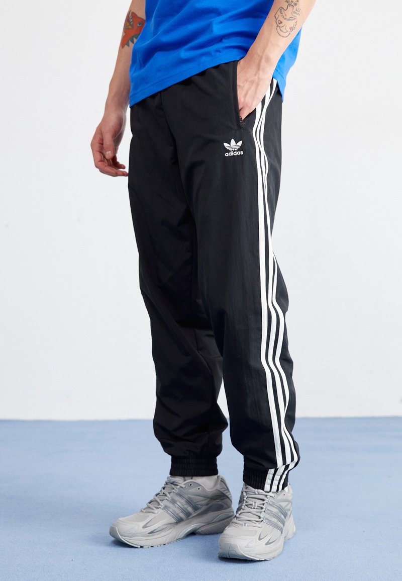 Svarta träningsbyxor med vita sidostrikar, elastiska muddarna och en Adidas-logotyp. Bärs med en blå skjorta och grå sneakers.