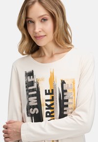 Koszula z długim rękawem w kremowym kolorze z nadrukiem z napisami "UŚMIECH", "BŁYSK" i "BLASK" ozdobionymi czarnymi i złotymi smugami farby.