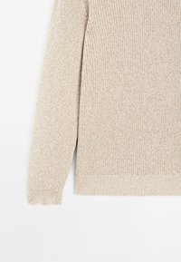 Manche de pull beige tricoté et détail sur le côté, montrant un tissu texturé et un poignet côtelé sur fond blanc.