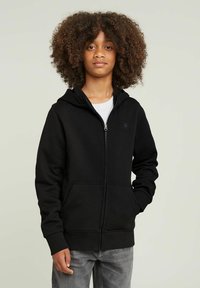 Sudadera negra con cremallera y bolsillo frontal, de textura suave y con puños acanalados. Llevada encima de una camiseta blanca con un logotipo sutil en el pecho.