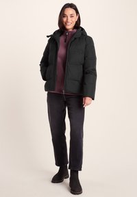 Smiling woman wearing black puffer jacket over purple zip-up sweater, dark jeans, and black boots, standing against plain light background.