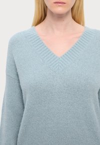 Hellblauer V-Ausschnitt-Pullover aus weichem, strukturiertem Stoff; verfügt über einen gerippten Kragen und überschnittene Schultern, die eine lockere Passform betonen.