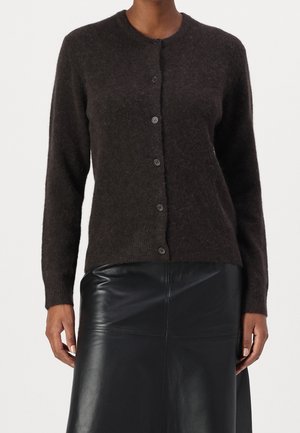 Femme portant un cardigan pelucheux marron foncé boutonné et une jupe en cuir noire, debout sur un fond clair uni.