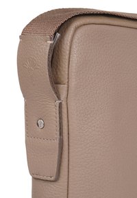 Angolo di una borsa in pelle marrone con finitura testurizzata e una tracolla intrecciata. Dotata di accessori in argento e dettaglio del logo impresso sulla tracolla.