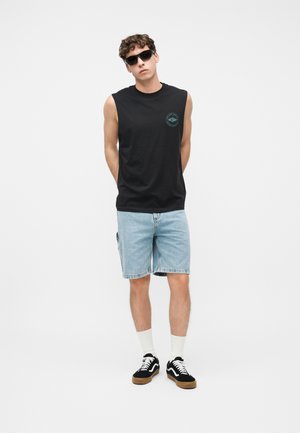 Joven con camiseta sin mangas negra, pantalones cortos de mezclilla claros, zapatillas negras con cordones blancos, calcetines blancos y gafas de sol negras, sobre fondo blanco.