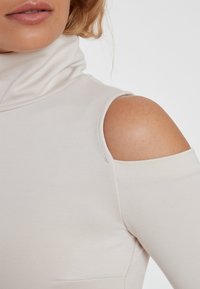Beige Rollkragenoberteil mit einem schulterfreien Design, das eine weiche, glatte Textur und eine figurbetonte Silhouette aufweist.