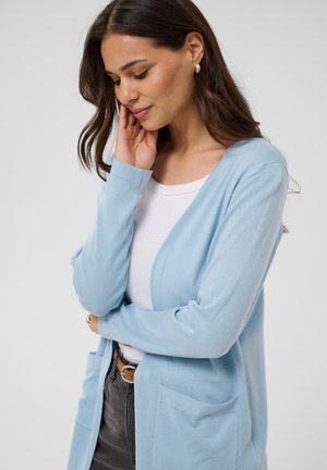 Femme aux longs cheveux bruns portant un cardigan bleu clair sur un haut blanc et un jean foncé, touchant doucement son visage d'une main.