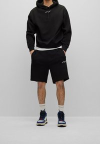 Sweat à capuche en coton noir avec poche avant et cordon de serrage, associé à un short noir assorti avec logo. Complété par des baskets bleues et blanches.