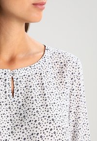 Blouse blanche à motif de points noirs, col rond, et petit détail en forme de goutte à l'avant. Légère, texture lisse.