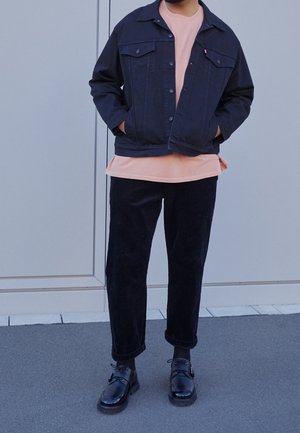 Personne portant une veste en denim noire sur un t-shirt rose, un pantalon noir, des chaussettes noires et des chaussures en cuir noires, debout contre un mur gris.