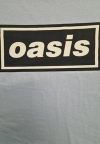 Lyseblå t-shirt med et fedt sort rektangulært grafisk design, hvor ordet "oasis" står med store hvide bogstaver centreret. Blødt stof.