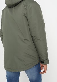 Grön vadderad jacka med huva, med en slät yta och elastiska muddar. Enkel design, sedd bakifrån, kombinerad med blå jeans.